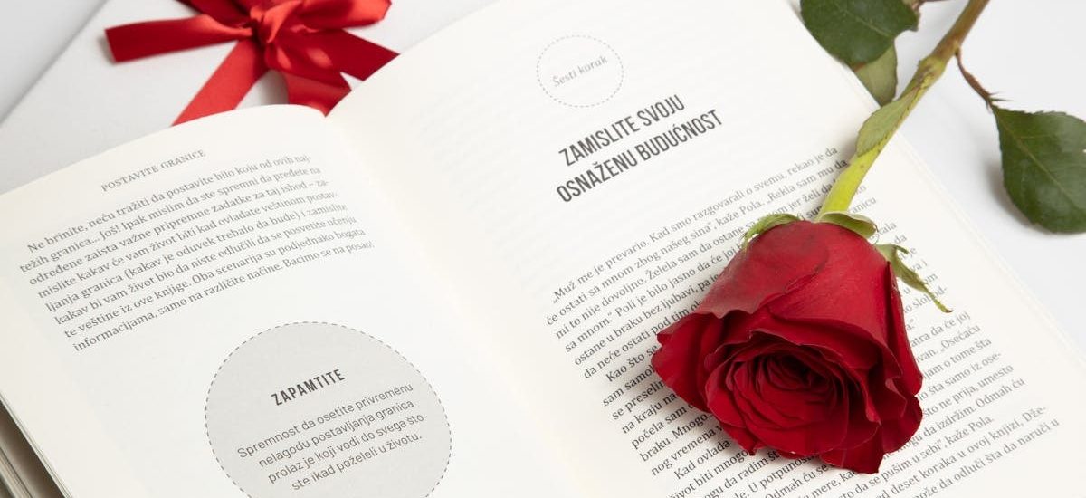 An open book with a red rose and a tied gift box next to it, conveying romance or celebration.